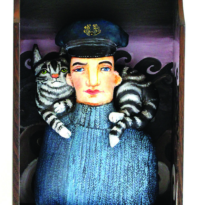 Harry McNish, dressed in a jumper and sailor cap, with his cat Mrs Chippy, on his shoulders. Fabric painted sculptures, inside a wood drawer. Rough sea waves are painted in the background.