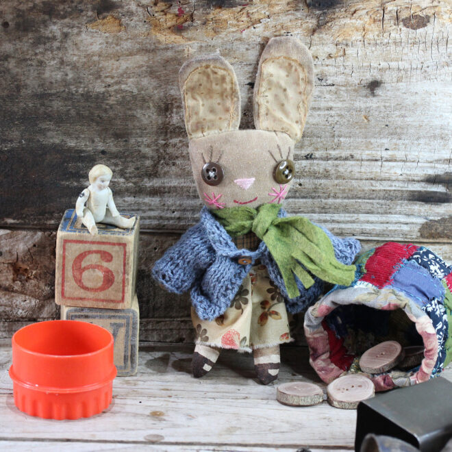 Small cloth doll in the form of an anthropomorphic bunny. She is wearing a denim coloured knitted caedigan, a cream and orange dress. Her face is embroidered and she has big brown buttons for eyes. Her shoes are hand painted. The cloth bunny has been distressed in coffee, giving her a vintage look.