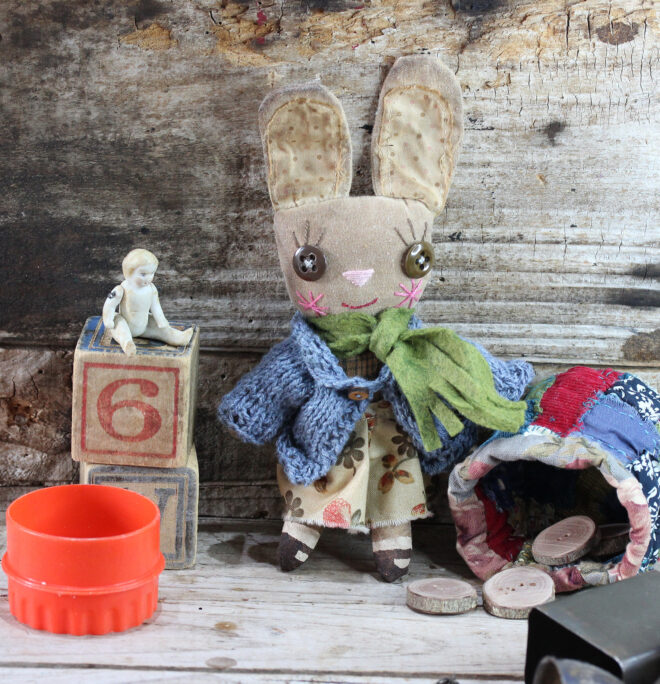 Small cloth doll in the form of an anthropomorphic bunny. She is wearing a denim coloured knitted caedigan, a cream and orange dress. Her face is embroidered and she has big brown buttons for eyes. Her shoes are hand painted. The cloth bunny has been distressed in coffee, giving her a vintage look.