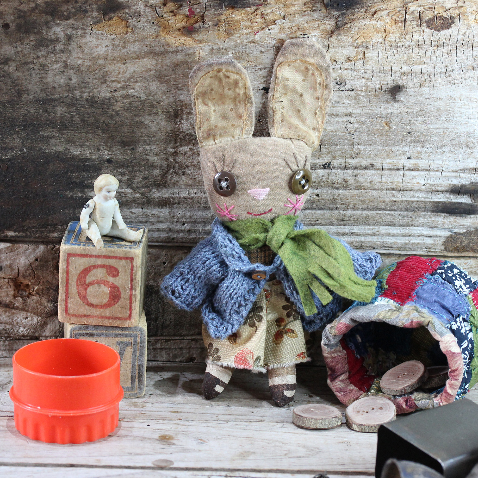 Small cloth doll in the form of an anthropomorphic bunny. She is wearing a denim coloured knitted caedigan, a cream and orange dress. Her face is embroidered and she has big brown buttons for eyes. Her shoes are hand painted. The cloth bunny has been distressed in coffee, giving her a vintage look.