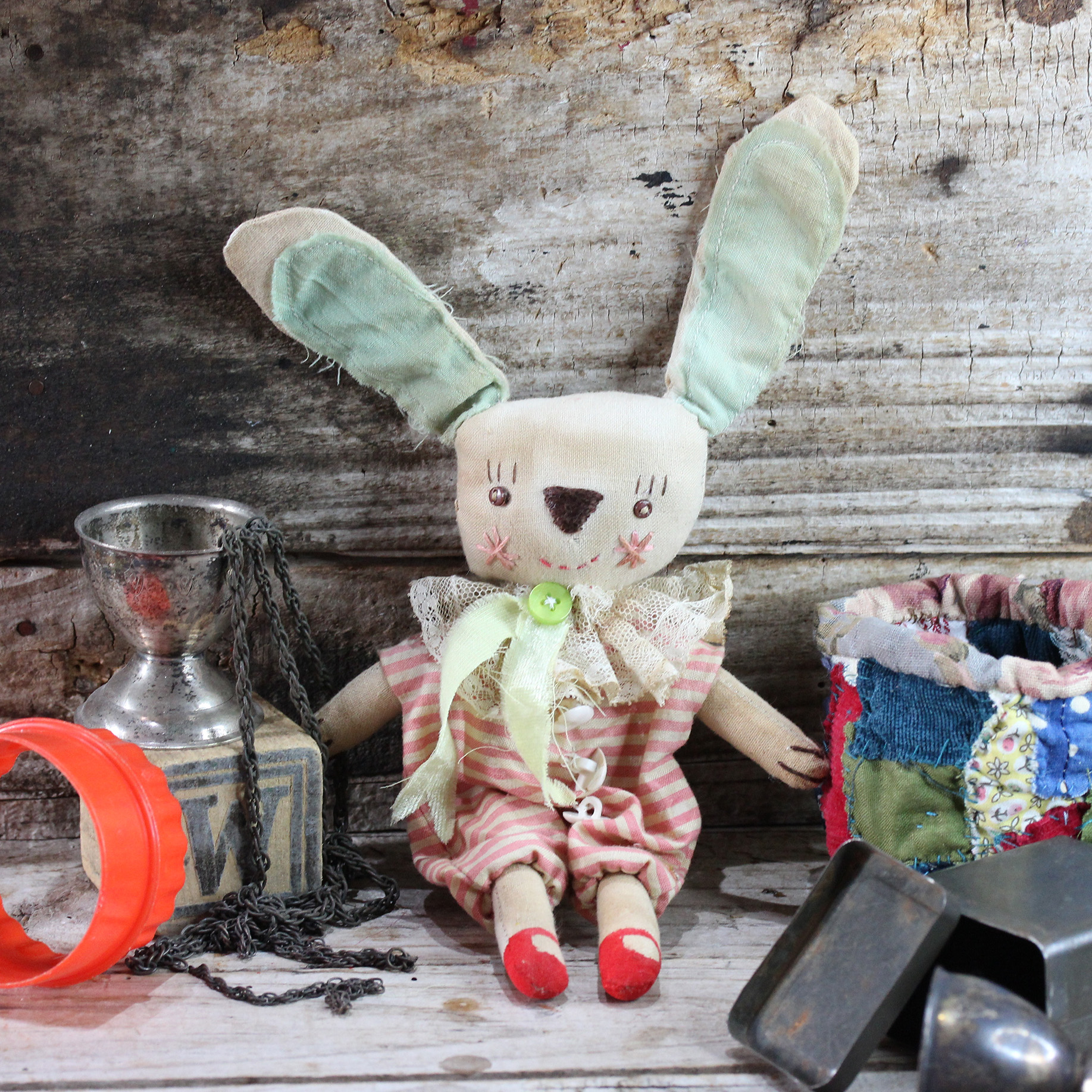A newly made cloth doll, stained to look old. Yhe anthropomorphic rabbit wears a pink and whit striped costume with a vintage lace collar. His face is embroidered with tiny black metal buttons for eyes.