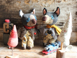 Picture shows two small Tasmanian devil dolls, wearing pants, cardigans and a wool felt scarf. Their cardigans are grey and their pants are yellow with woodland patterns. The devils are painted black with brown snouts.