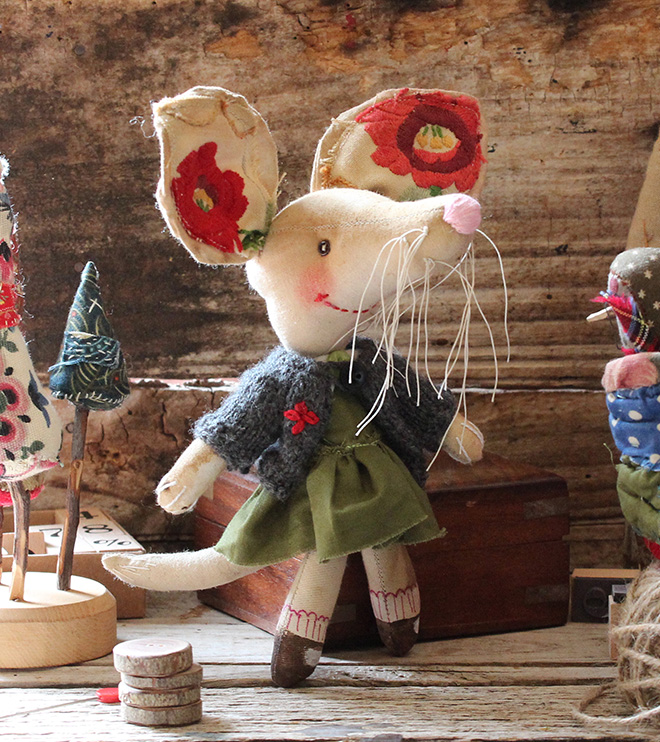 Coffee stained anthropomorphic small calico doll in the form of a Bilby. She wears a green dress and grey cardigan, red shoes and striped socks. Her ears are large and embroidered, her whiskers are long and white, eyes are tiny buttons. Her moth is hand stitched and she has rosey cheeks.