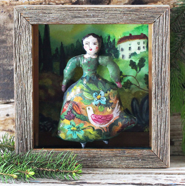 Inside a small shadow box is a fabric art doll wearing a green painted, stylized Victorian dress with birds and flowers decorated in the skirt.